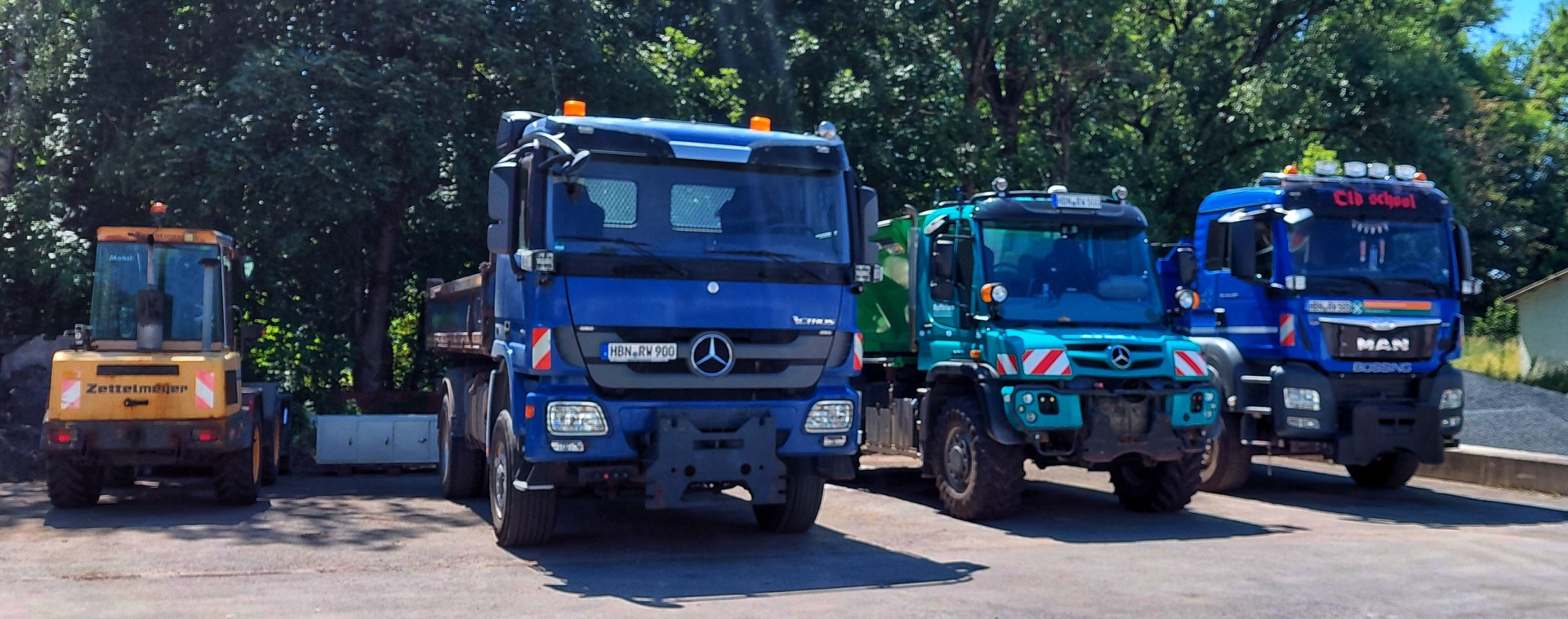 Line of heavy construction trucks parked on a sunny lot, including a yellow excavator and several blue Mercedes trucks with trees in the background.