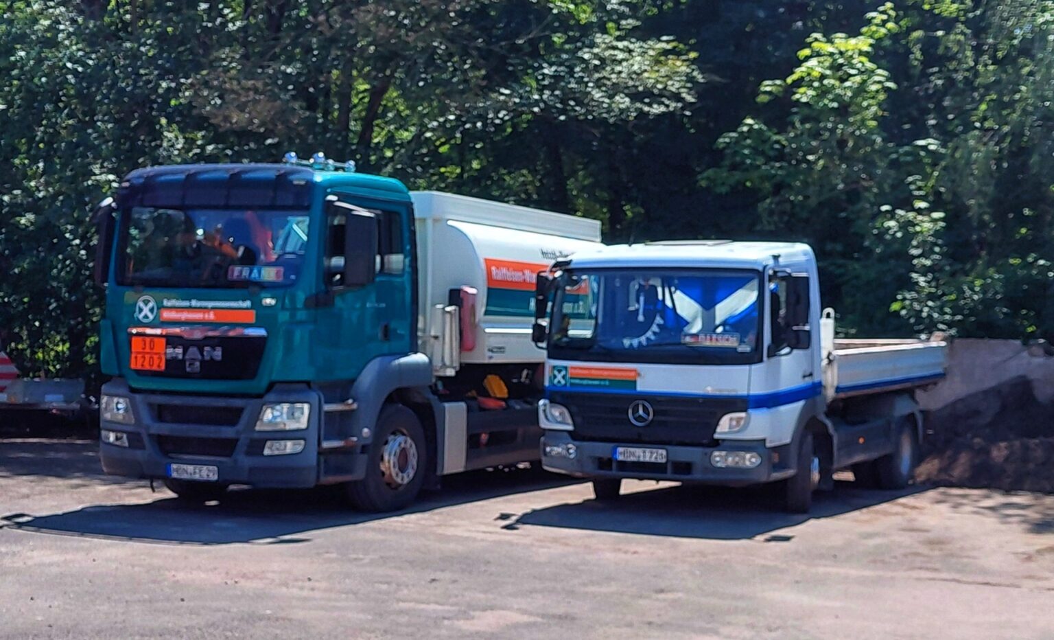 Two large trucks parked side by side on a road, a green truck in front and a white truck behind, with trees in the background.