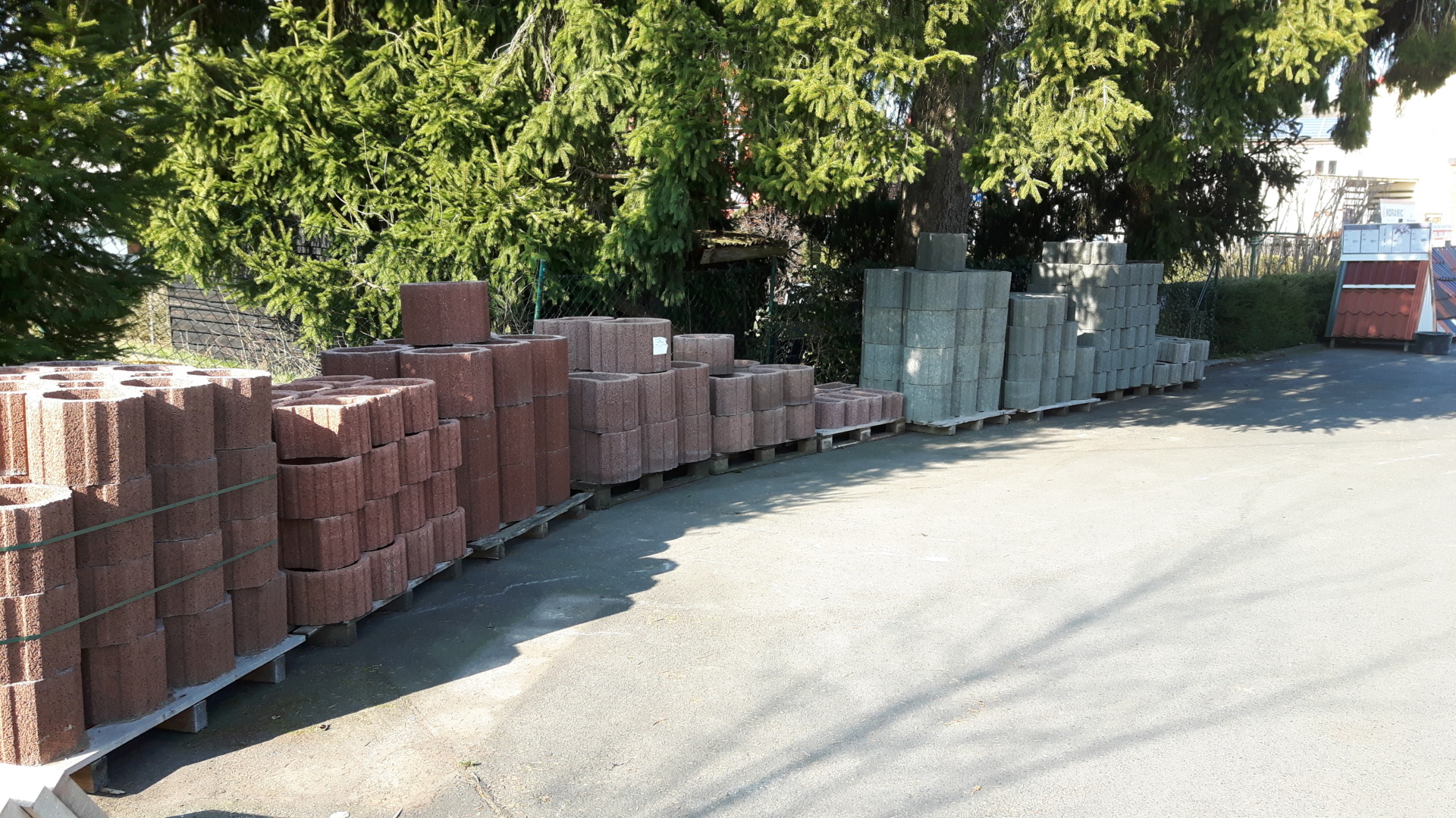 Stacks of red and gray concrete pavers on pallets along a road, with evergreen trees and a fence in the background.